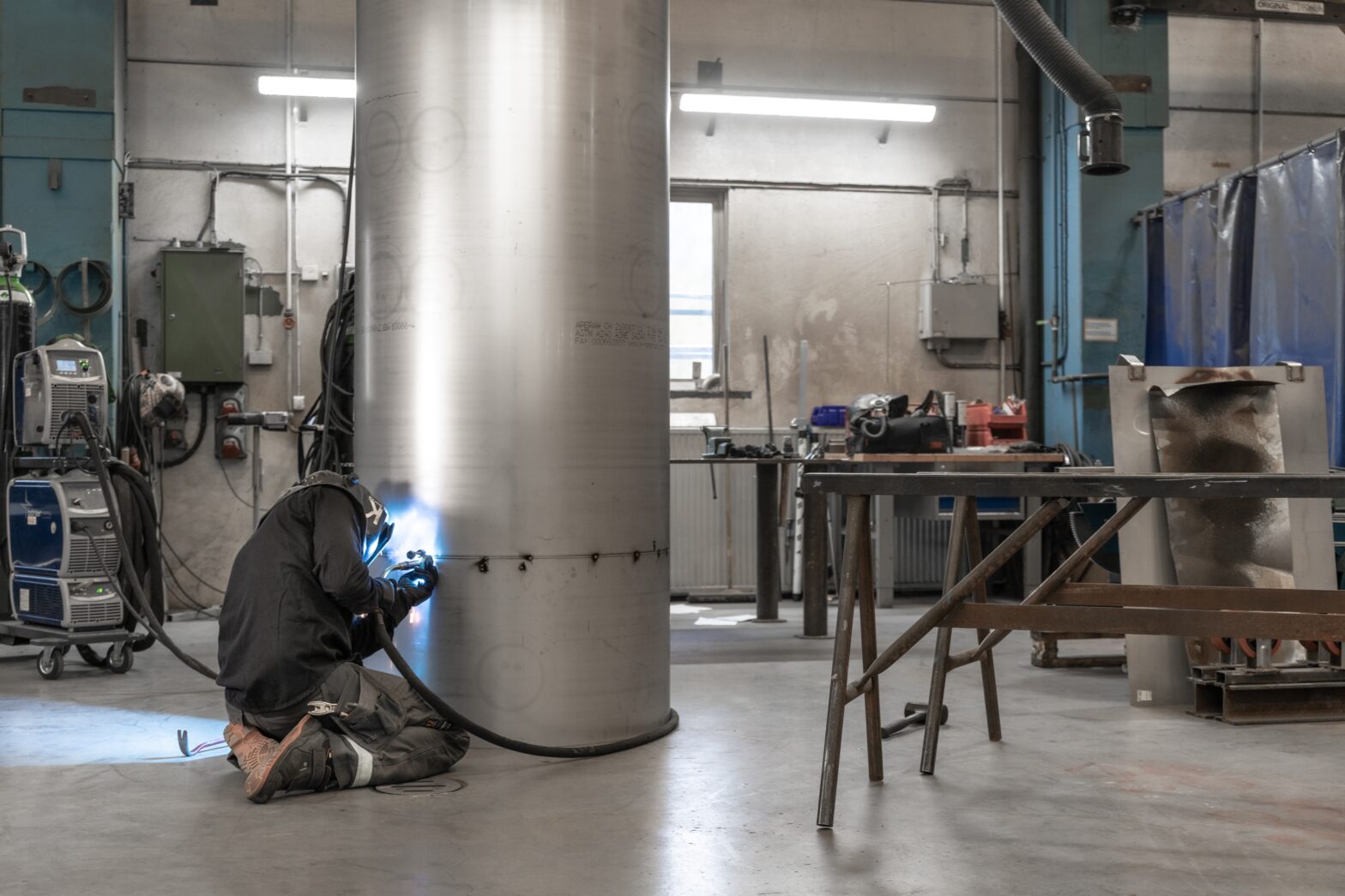 An image of a workshop, with a person welding on a floor to ceiling length aluminium cylinder