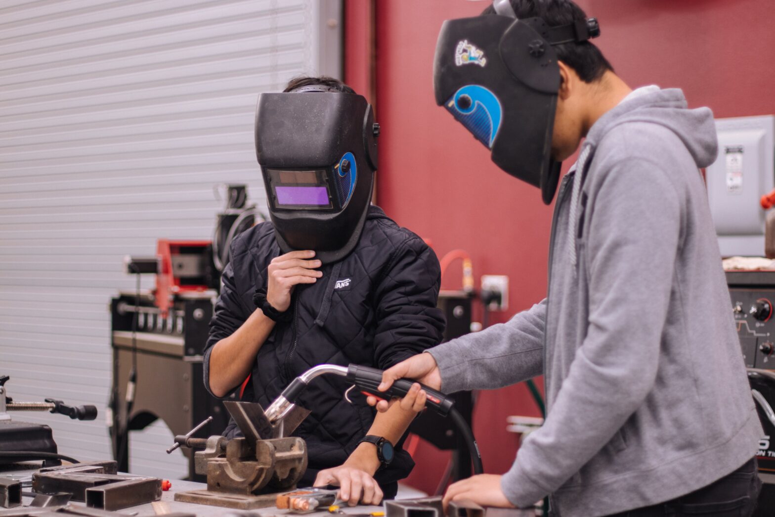 A photograph of two people in a workshop, using welding equipment to weld materials. They are both wearing helmets and the correct welding PPE.