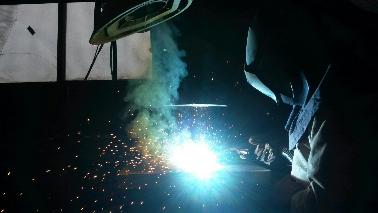 A welder working with intense blue light emitting from the welding spot, creating a shower of bright orange sparks, while smoke rises around the area, all taking place in a dimly lit workshop